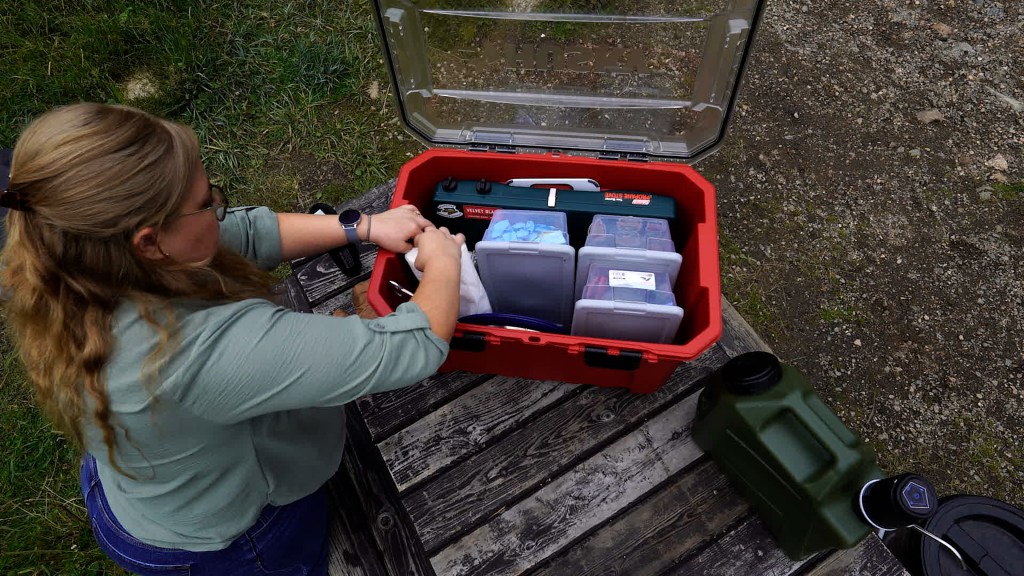 A person organizing a red camp kitchen tote filled with various containers and kitchen supplies, set on a wooden surface outdoors.