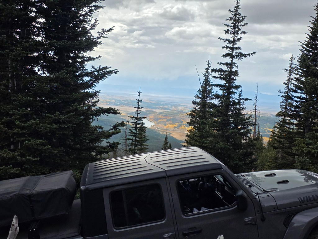 A panoramic view from a mountain overlook showing a valley with colorful autumn foliage, framed by evergreen trees and a parked grey Jeep Gladiator vehicle.