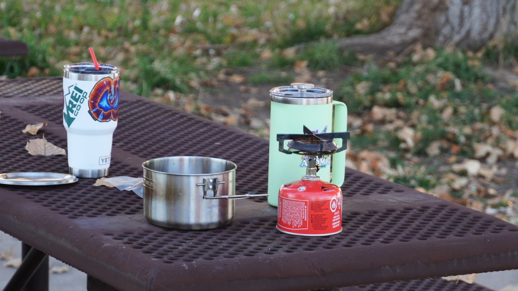 A portable camping stove with a red propane canister, a stainless steel cooking pot, and a green insulated YETI French Press coffee drink container are set on a picnic table covered with fallen leaves.