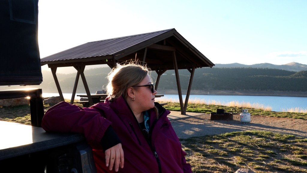 A person in a maroon jacket sitting by the water with a scenic view of a lake and mountains in the background. The sun is shining, casting a warm glow on the scene.