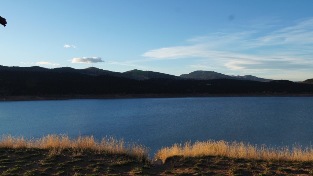 A serene view of a tranquil reservoir surrounded by rolling hills and distant mountains under a clear blue sky, with tall grasses lining the shore.