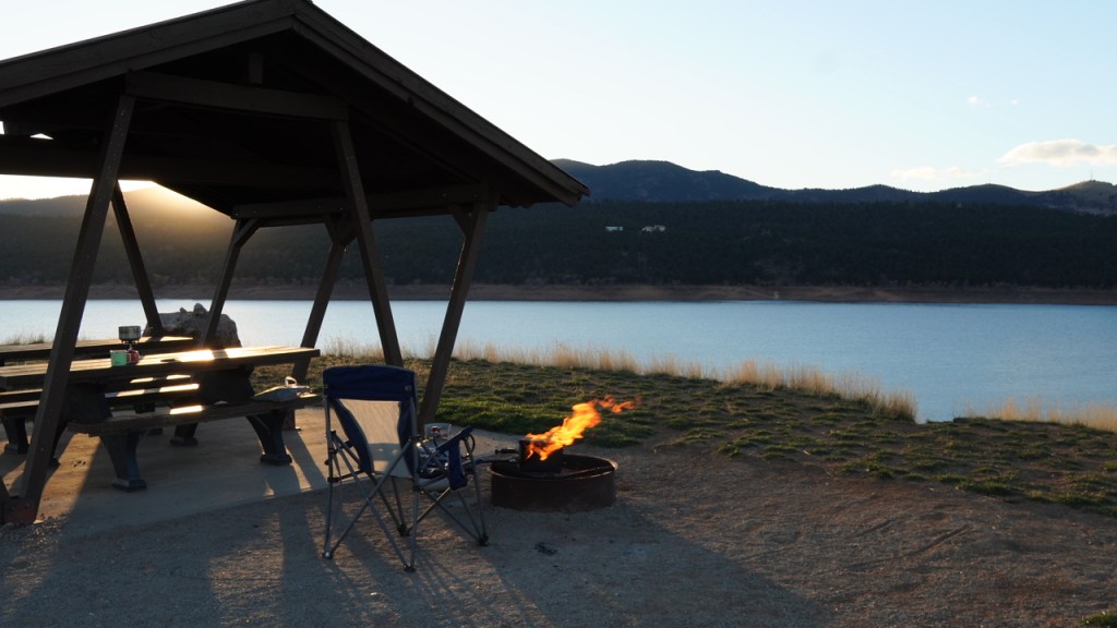 A peaceful picnic shelter with a fire pit near a lake, mountains in the background, and sunlight casting shadows on the ground.
