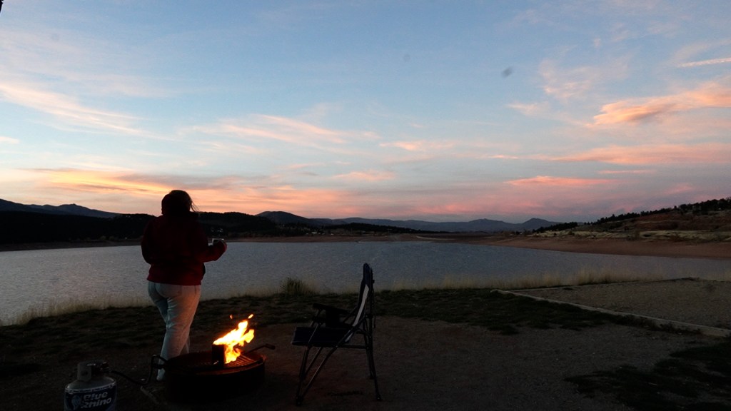 A person standing near a fire pit by a lake at sunset, with mountains in the background and colorful clouds in the sky.