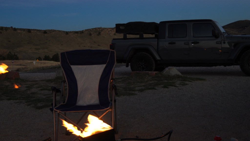 A camp chair next to a portable fire pit with flames, set against a twilight landscape featuring a Jeep truck and distant hills.