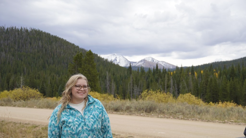 A person standing along a dirt road in a mountainous area, surrounded by evergreen trees and a backdrop of snow-capped peaks under a cloudy sky.