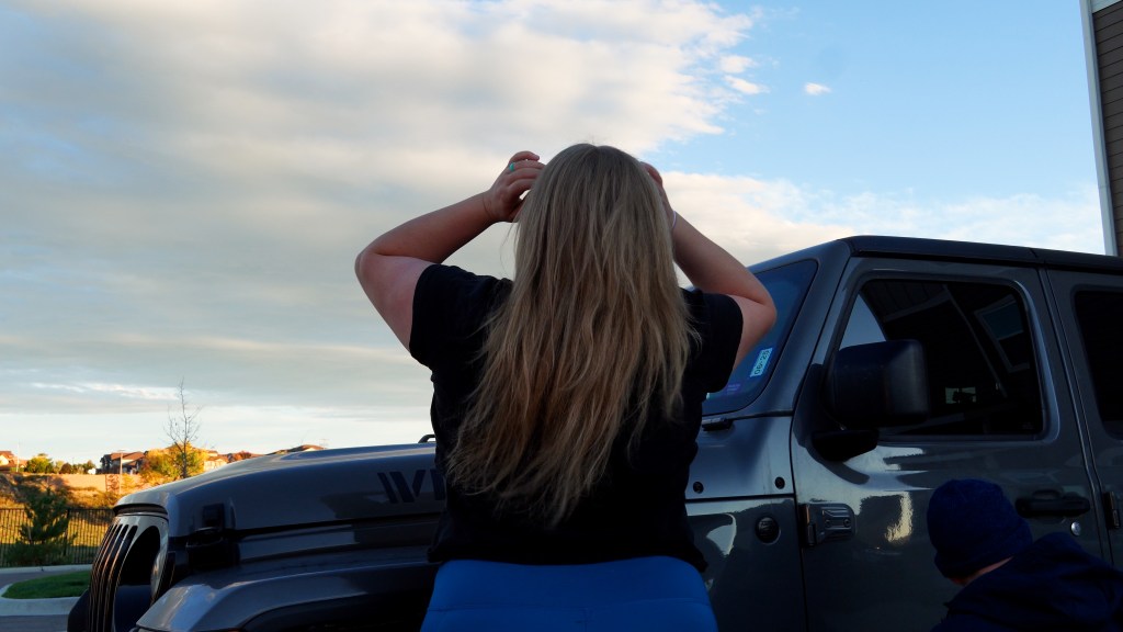A person with long hair stands behind a gray jeep gladiator, looking up at the sky with clouds, while another person crouches nearby.